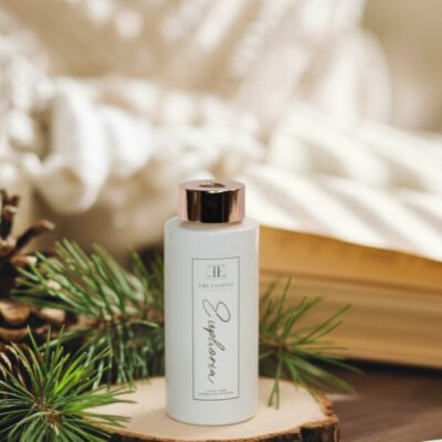A white reed diffuser and resting on a wooden slice, surrounded by pinecones, pine needles, and scattering of pine nuts on a dark wooden table.